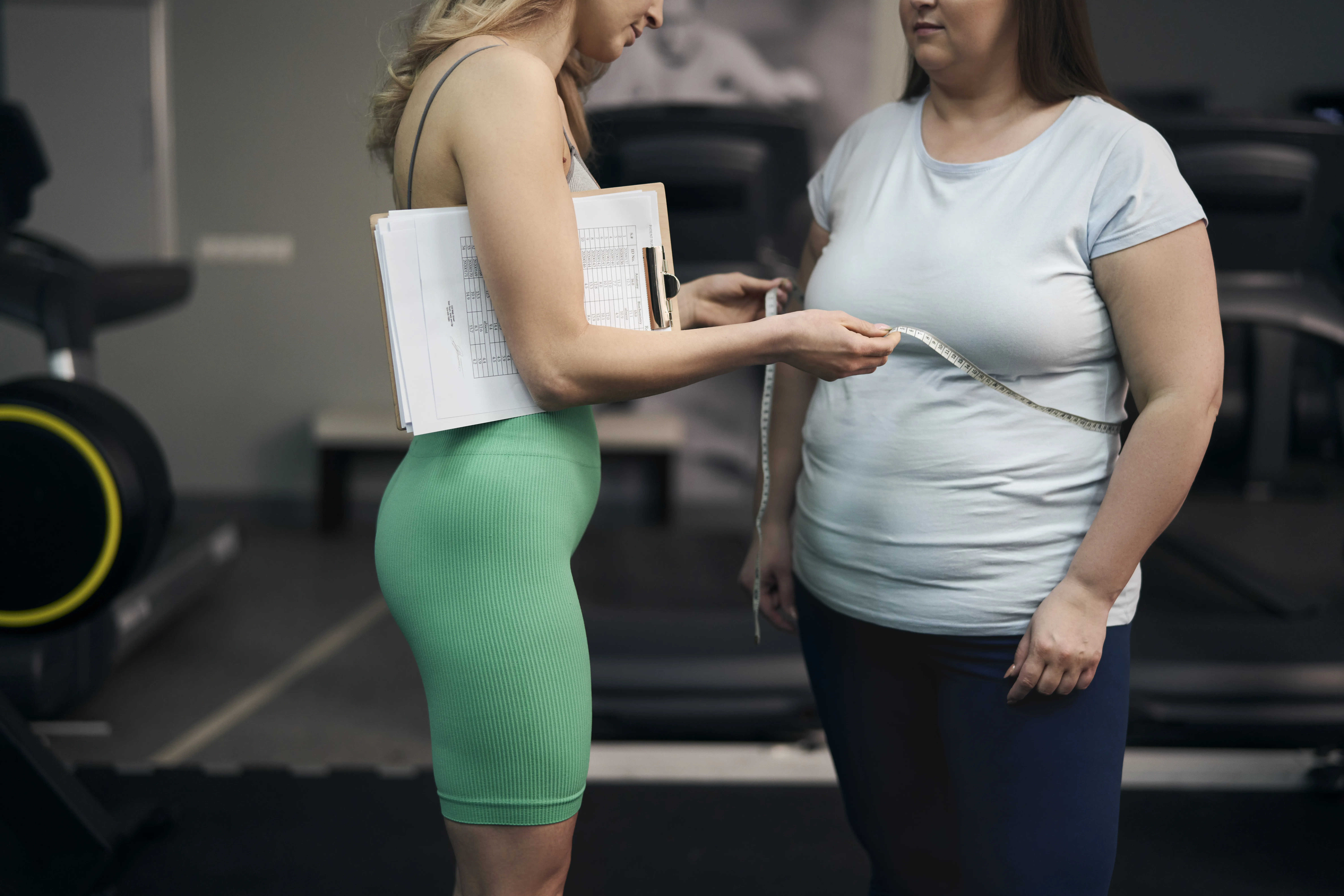 Trenerka lub dietetyczka w zielonym stroju sportowym mierzy centymetrem obwód talii pacjentki na siłowni, trzymając pod pachą podkładkę z wynikami pomiarów.